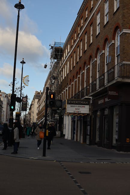 A street scene on Baker Street in Marylebone shows a row of multi-storey brick buildings with arch-shaped windows and small balconies. Several pedestrians are walking along the pavement, some carrying bags. Traffic lights are visible, with one showing a red pedestrian signal. A sign indicates the location as Baker Street Moves, offering house removals, and is attached to a storefront. Above, scaffolding covered with protective netting is fitted to the side of a building under construction. Street lamps line the road, and a sky with a few clouds is partly visible overhead. The environment suggests an active urban area suitable for home relocation and furniture transport activities, with the company Man with Van Marylebone operating in the vicinity, supporting moving and packing logistics. The scene captures a typical busy day in Marylebone during a furniture transport or loading process for house removals.