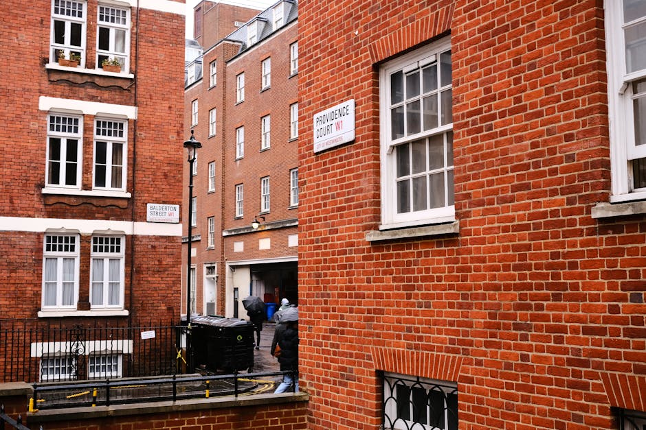 A busy street scene in Marylebone with a large multi-storey brick and white building on the corner, featuring several windows and a ground-floor retail space. The retail area includes signage and window displays, with pedestrians crossing the road at a marked pedestrian crossing. Some individuals are carrying shopping bags or backpacks, and others are walking along the sidewalk. Vehicles, including a small white car, are waiting at the traffic lights. Street furniture such as traffic lights, street lamps, and a signpost indicating 'Baker Street NW1' are visible. The area appears well-lit with natural daylight, and the scene captures a typical urban environment suitable for home relocation and furniture transport services, like those provided by Man with Van Marylebone, supporting logistics and packing during house removals in the Marylebone area.