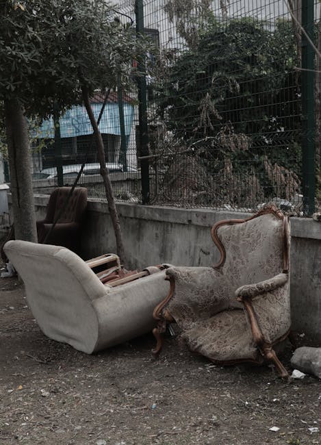 A pair of vintage armchairs and a small sofa with wooden frames and fabric upholstery, all showing signs of weathering and dirt, are positioned outdoors on a gravel surface near a concrete wall and metal fence. The armchairs have ornate, curved wooden armrests and high backs, with the upholstery appearing faded and worn. The small sofa has a simpler design with a rounded back and wooden legs, with its fabric visibly stained and sagging. Behind the furniture, there are trees and foliage providing a background, with some parts of the outdoor setting partially obscured by leaves and branches. The scene appears to be part of a collection of discarded furniture outside, possibly for removal or disposal, and the area is illuminated by natural daylight. This image aligns with concepts of furniture disposal and home clearance, as handled by services such as Man with Van Marylebone.