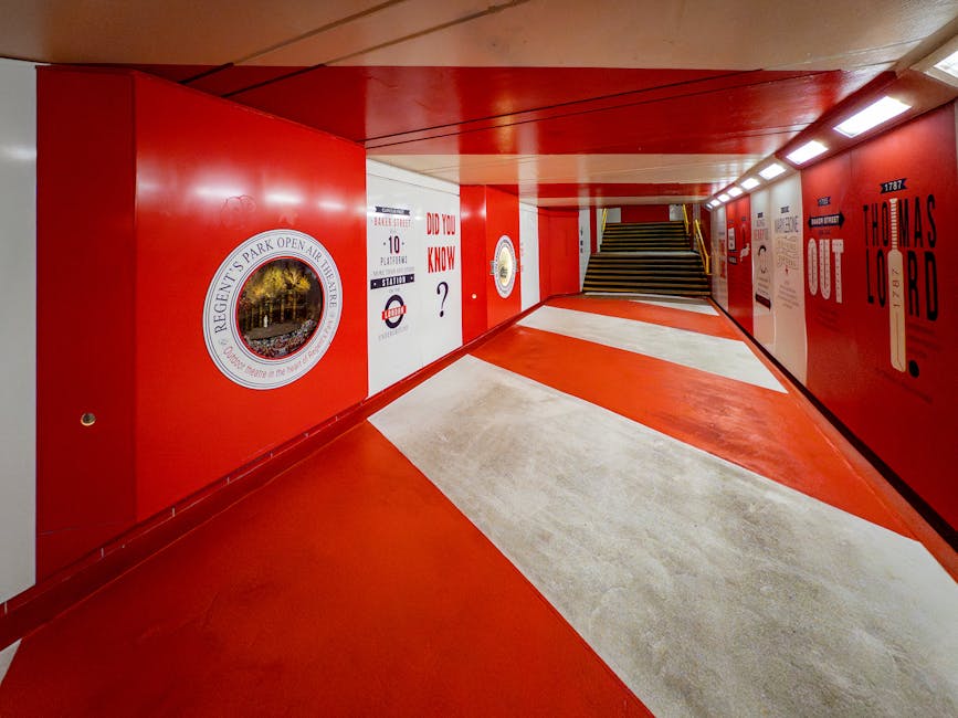 The image shows an underground pedestrian tunnel with bright red walls and a matching red ceiling, illuminated by long, white LED lights. The floor features a distinctive pattern of red and cream-colored sections leading towards a staircase at the far end, which appears to be part of a home relocation or building access point. The tunnel walls display various posters, signs, and markings, including a large circular emblem with 'Regent's Park Open Air Theatre' and other smaller informational notices, some referencing London transportation and history. The staircase leading upstairs has a black and yellow safety strip along the edge of each step. The environment looks clean and well-maintained, suggestive of a location where furniture transport, packing, and moving logistics may be coordinated, reflecting services offered by Man with Van Marylebone for efficient local moves in the Marylebone area.