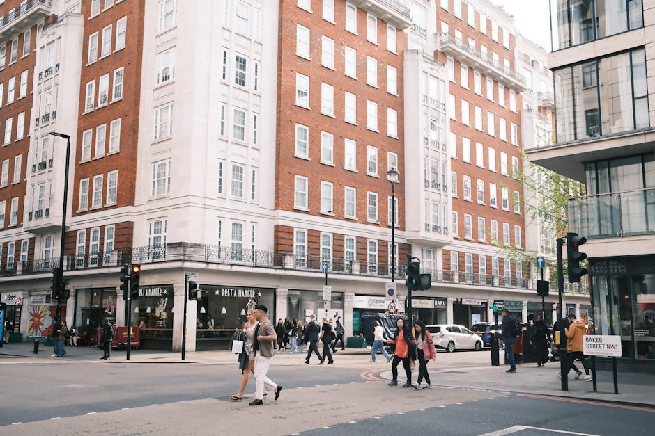 A busy urban street scene at a crossroad in Marylebone, featuring a large multi-storey red brick and white façade building with retail shops on the ground floor, including a Pret A Manger and other stores, with pedestrians crossing the street and walking along the sidewalk. Several people carry shopping bags or backpacks, and some are engaged in conversation. Parked and moving vehicles, including a white car, are visible along the curb, and black traffic lights and street lamps are positioned at the intersection. The street is paved with asphalt, with pedestrian crossings marked on the road. The scene is well-lit with natural daylight, capturing a typical city environment suitable for the content on the Marylebone High Street Removals page associated with Man with Van Marylebone, which offers house removals and furniture transport services.