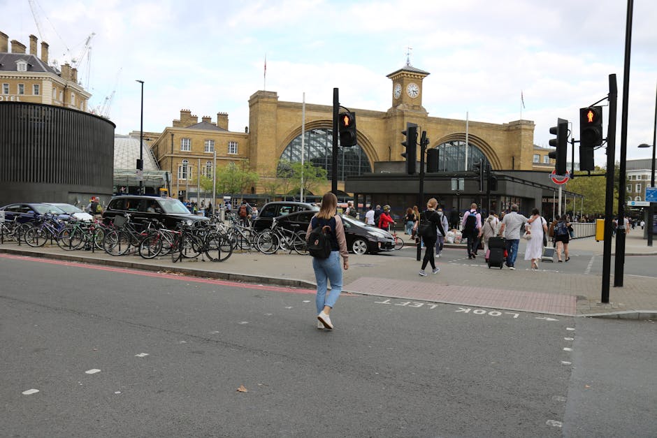 A busy street scene outside Marylebone Station in London, with several pedestrians crossing at a designated crossing area marked 'LOOK LEFT' on the pavement. In the background, the station's architectural facade features large arched windows and a clock tower, with a mix of historic and modern buildings nearby. On the left, a row of bicycles is secured to bike racks, and nearby parked cars are visible. The street is lined with traffic lights; some are showing red signals for vehicles. The lighting suggests an overcast day. This setting exemplifies the urban environment where professional house removals or furniture transport services, provided by companies like Man with Van Marylebone, often coordinate loading and unloading operations, including parking and meet-up points close to the station for efficient home relocation processes.