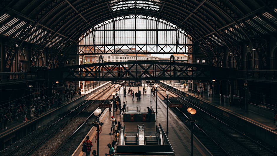 Inside Marylebone Station in London, an expansive train station with high arched metal and glass roofing allowing natural light to fill the space. The station features multiple train tracks with platforms on each side, crowded with passengers waiting or walking, some carrying luggage or bags. In the foreground, there are several cardboard boxes, wrapped with plastic and placed on dollies, possibly ready for home relocation or furniture transport. A few flat-pack furniture pieces and moving blankets are visible among the boxes, indicating the process of packing and moving services. The station’s interior includes vintage-style lamp posts and signage, with passengers walking towards the trains or exits, and a staircase and escalator nearby for access to different levels. The overall scene captures the busy loading and unloading environment typical of a house removal process in progress at a major London transport hub, with the company Man with Van Marylebone occasionally mentioned for legal and service context, aligned with removals and moving logistics.