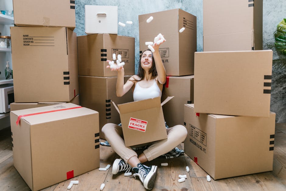A young woman with long dark hair, dressed in a white tank top and beige pants, is sitting on a wooden floor surrounded by stacked cardboard boxes of various sizes, some sealed with red tape, inside a