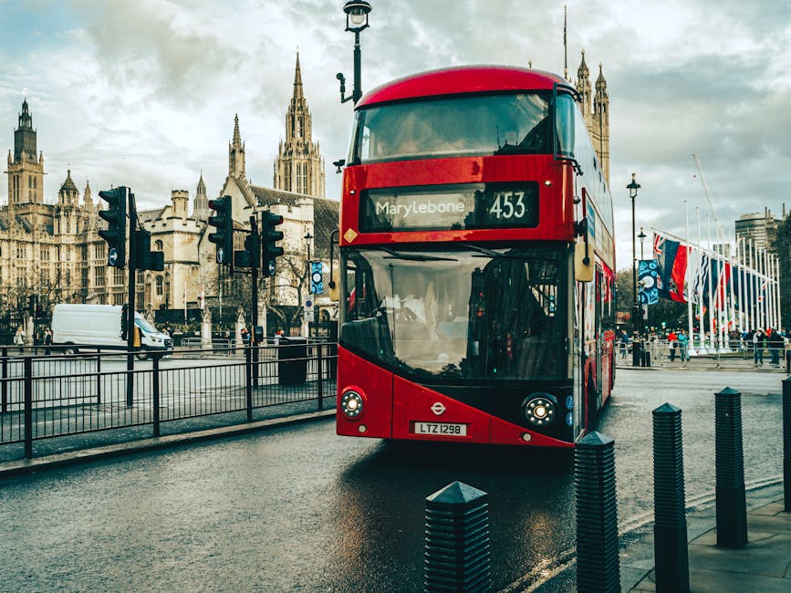 A red double-decker bus displaying the route number and destination as 'Marylebone' approaches a street in central London during daytime, with historic Gothic-style buildings and a cloudy sky in the background. The bus is in motion on a wet road, bordered by metal bollards and a pedestrian pathway, with traffic lights and a white van visible nearby. On the right side, a row of tall flagpoles with colorful flags is seen, and pedestrians are walking along the pavement, indicating an active urban environment. The scene depicts the typical setting for a home relocation process involving furniture transport, with the bus likely being part of local transportation services by Man with Van Marylebone, supporting efficient moving and packing logistics in the area.