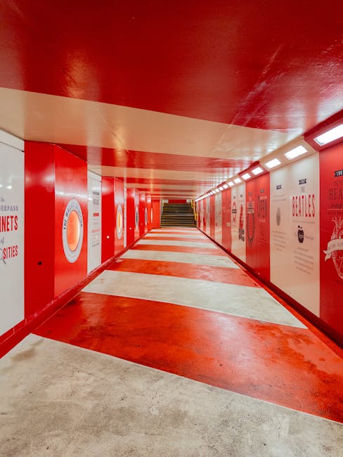 A spacious underground pedestrian walkway with a predominantly red colour scheme, featuring red-painted walls and ceiling, and a patterned floor with alternating red and white sections. Along the right side of the corridor, there are illuminated rectangular light fixtures and a series of posters or informational signs attached to the wall. Visible in the background are stairs leading upwards, indicating an exit or connection to other floors. The environment appears clean and well-maintained, designed for foot traffic, and the image context aligns with the type of access challenges faced during home relocations or furniture transport in tight or restricted staircase areas, as addressed by Man with Van Marylebone's removal services.