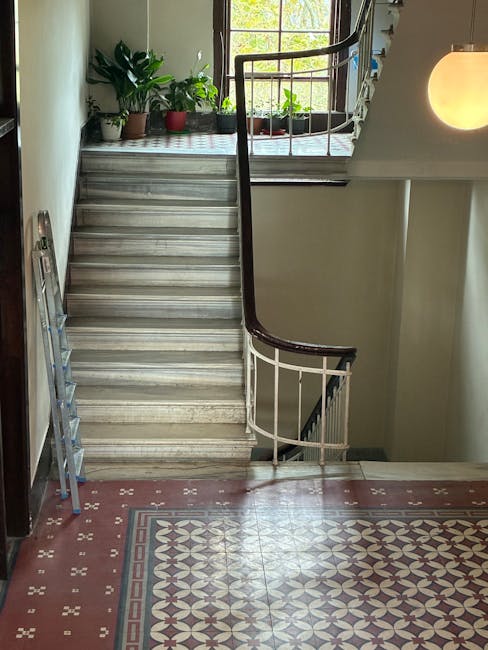 Inside a residential building showing a staircase with marble steps and a dark wooden handrail, leading upward to a landing with three potted plants near a window. The window provides natural light, illuminating the area and revealing a glimpse of greenery outside. A silver, multi-step step ladder is leaning against the left wall near the bottom of the stairs, suggesting ongoing or planned adjustments related to house moving or packing tasks. Adjacent to the staircase is a patterned tile floor in red and cream tones, with a round, warm-toned wall-mounted light fixture casting soft illumination on the wall. The environment indicates a typical home interior, aligned with furniture transport and home relocation services provided by Man with Van Marylebone, and highlights potential staircase access challenges common during furniture transport in such properties.