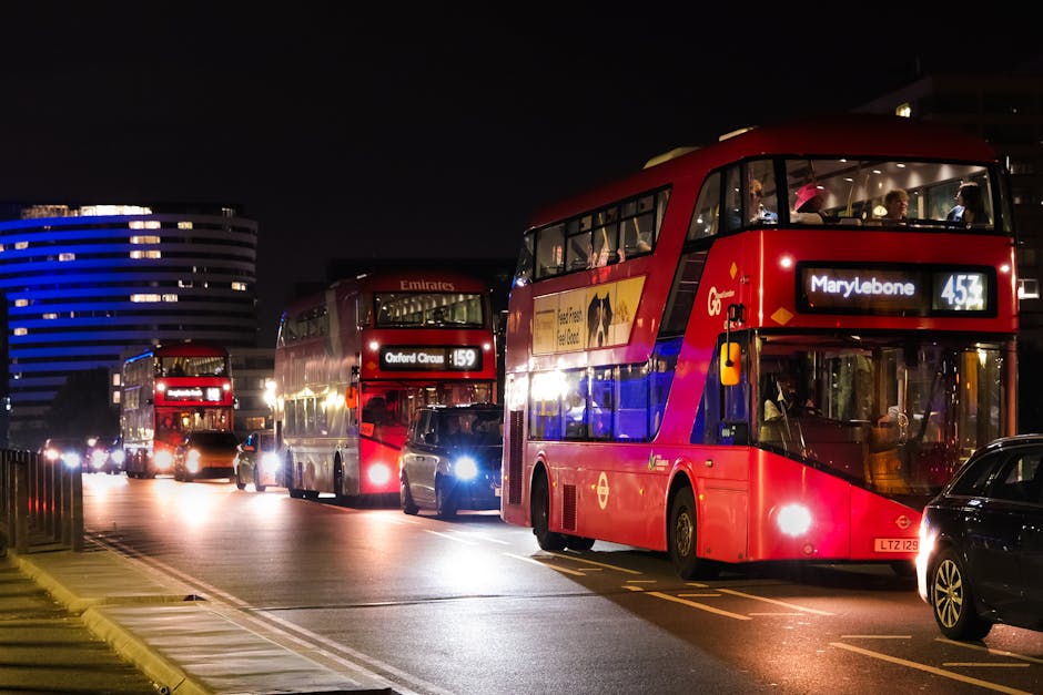 Nighttime scene in central London showing multiple red double-decker buses, including one with a sign indicating route 453 to Marylebone, parked on the street near the pavement. Behind the buses, a modern office building with illuminated blue horizontal lines is visible against a dark sky. The buses are positioned in a queue, with some headlights and interior lights turned on, reflecting off nearby vehicles and the road surface. The foreground includes a black car partially visible on the right side, parked in the street. The scene suggests a busy urban environment, possibly during evening hours, with ongoing transit operations related to city transport services such as those managed by companies like Man with Van Marylebone for house removals and moving logistics.